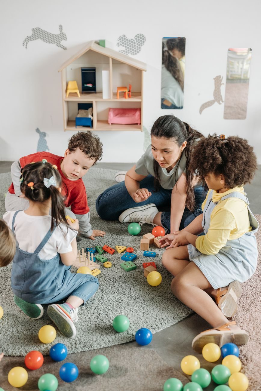 A teacher sitting on the carpet with four toddlers, sorting colored wooden shapes beside a wall-mounted dollhouse.