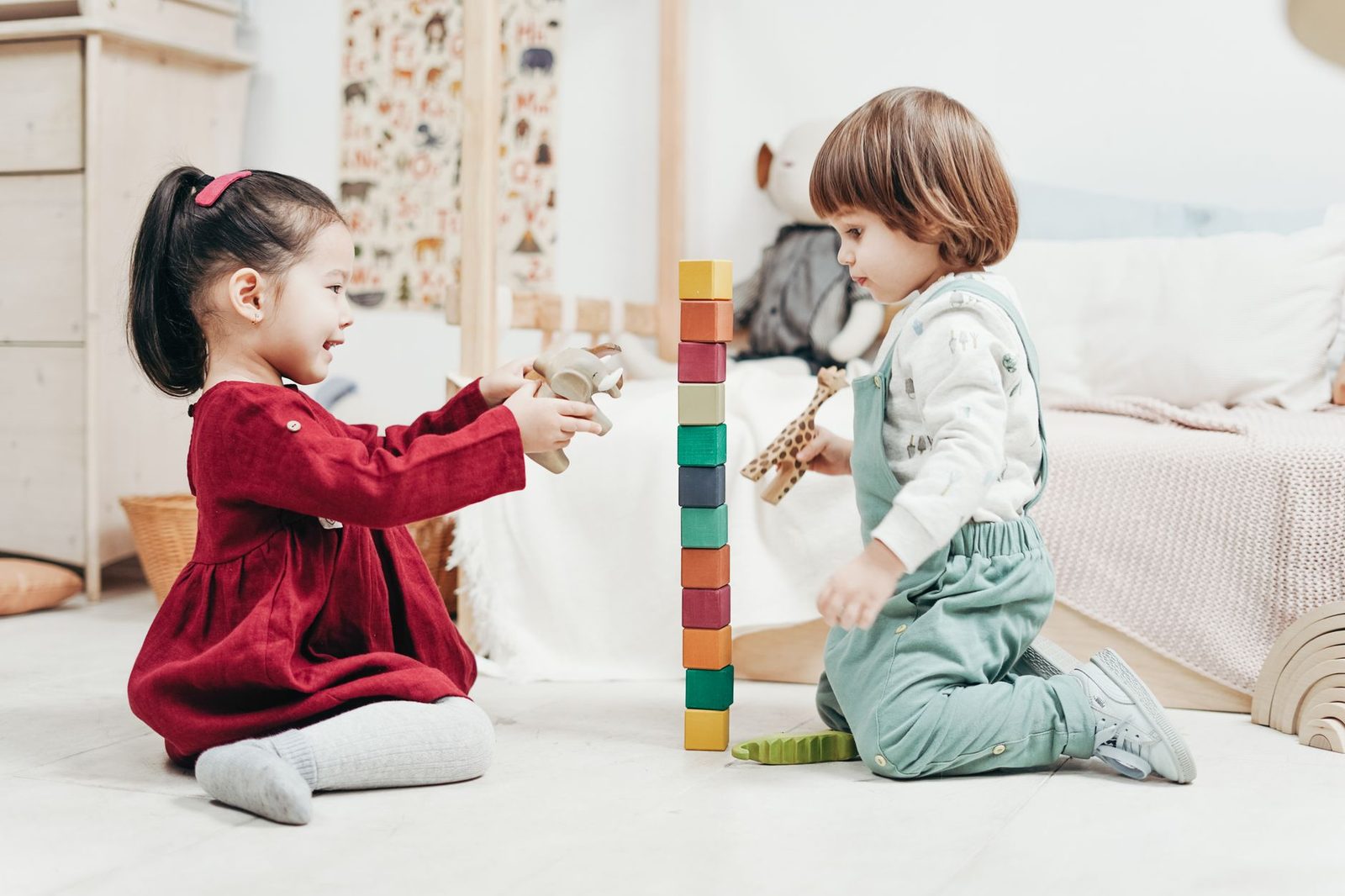 Two toddlers sitting facing each other on a sunlit floor, building a tall stack of colored wooden blocks between them.