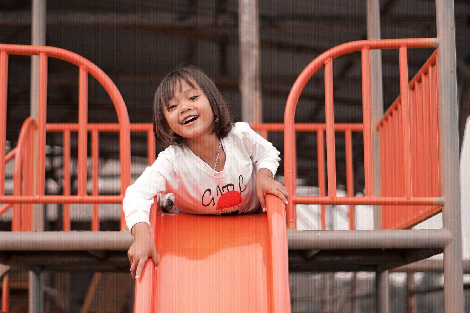 A grinning child at the top of a bright orange playground slide, hands on the rails, about to push off.