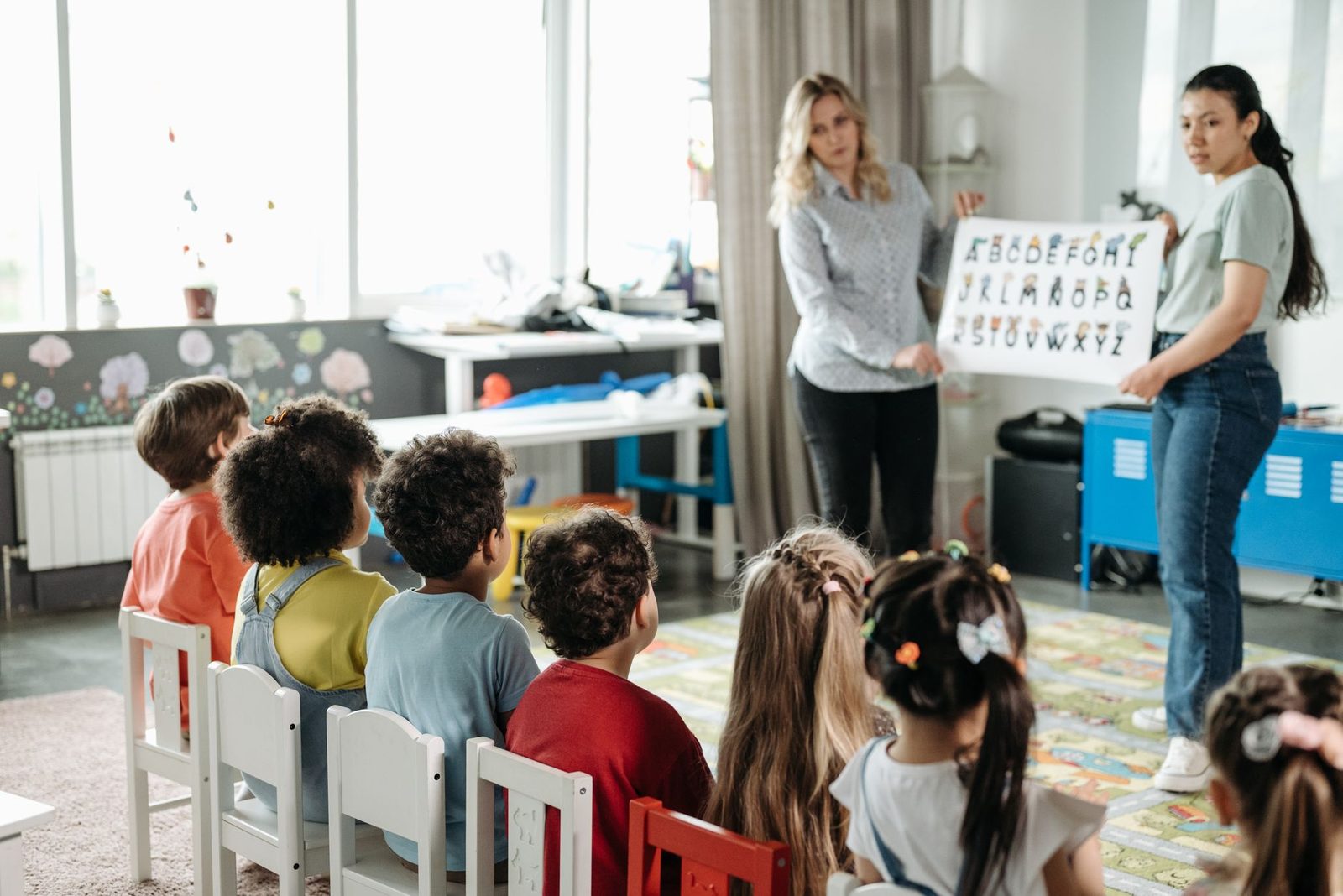 Two teachers holding up an alphabet chart for a class of children sitting on small chairs in a bright room.