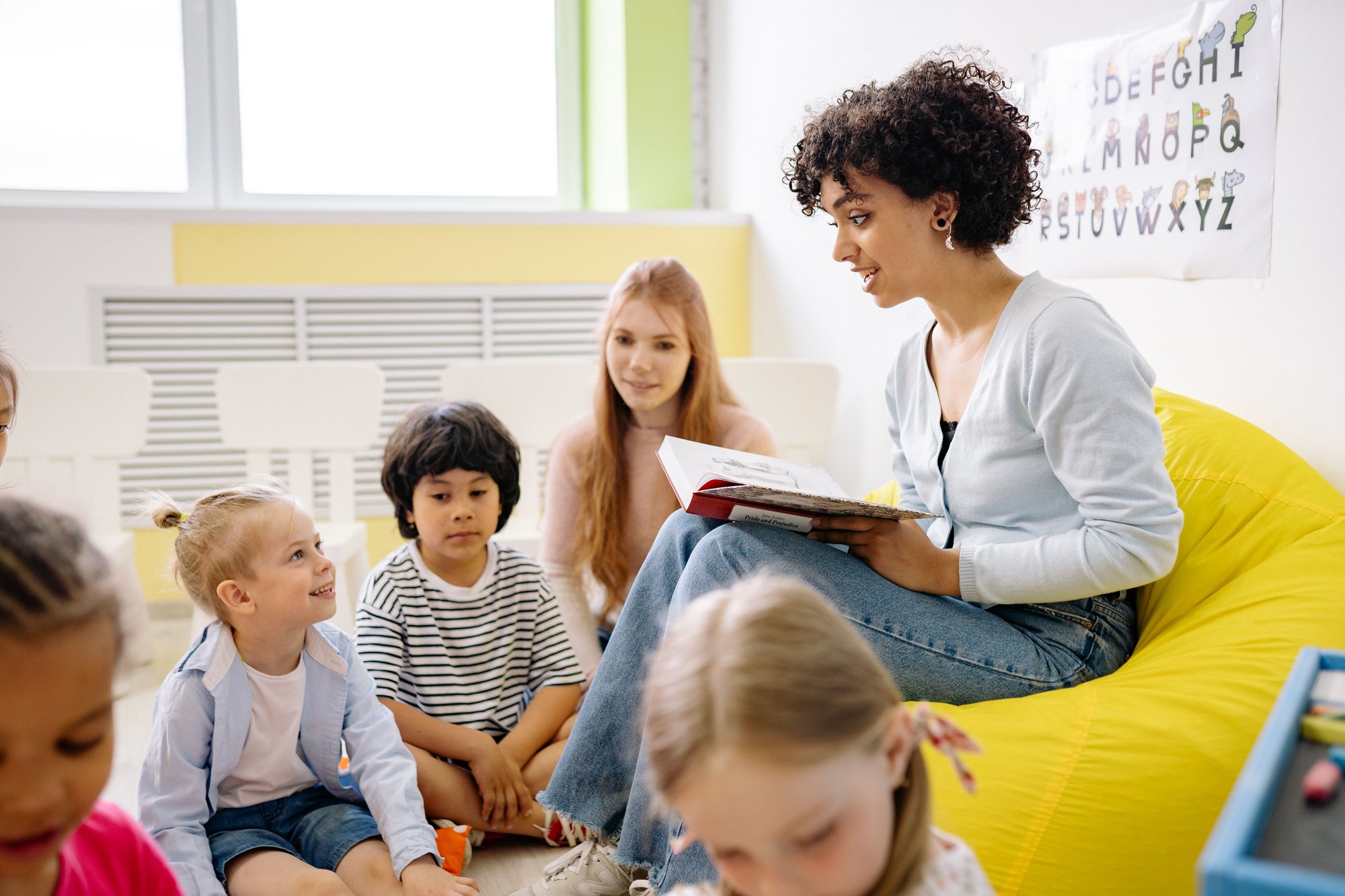 A teacher on a yellow beanbag reading a picture book to five preschoolers sitting cross-legged on the floor.