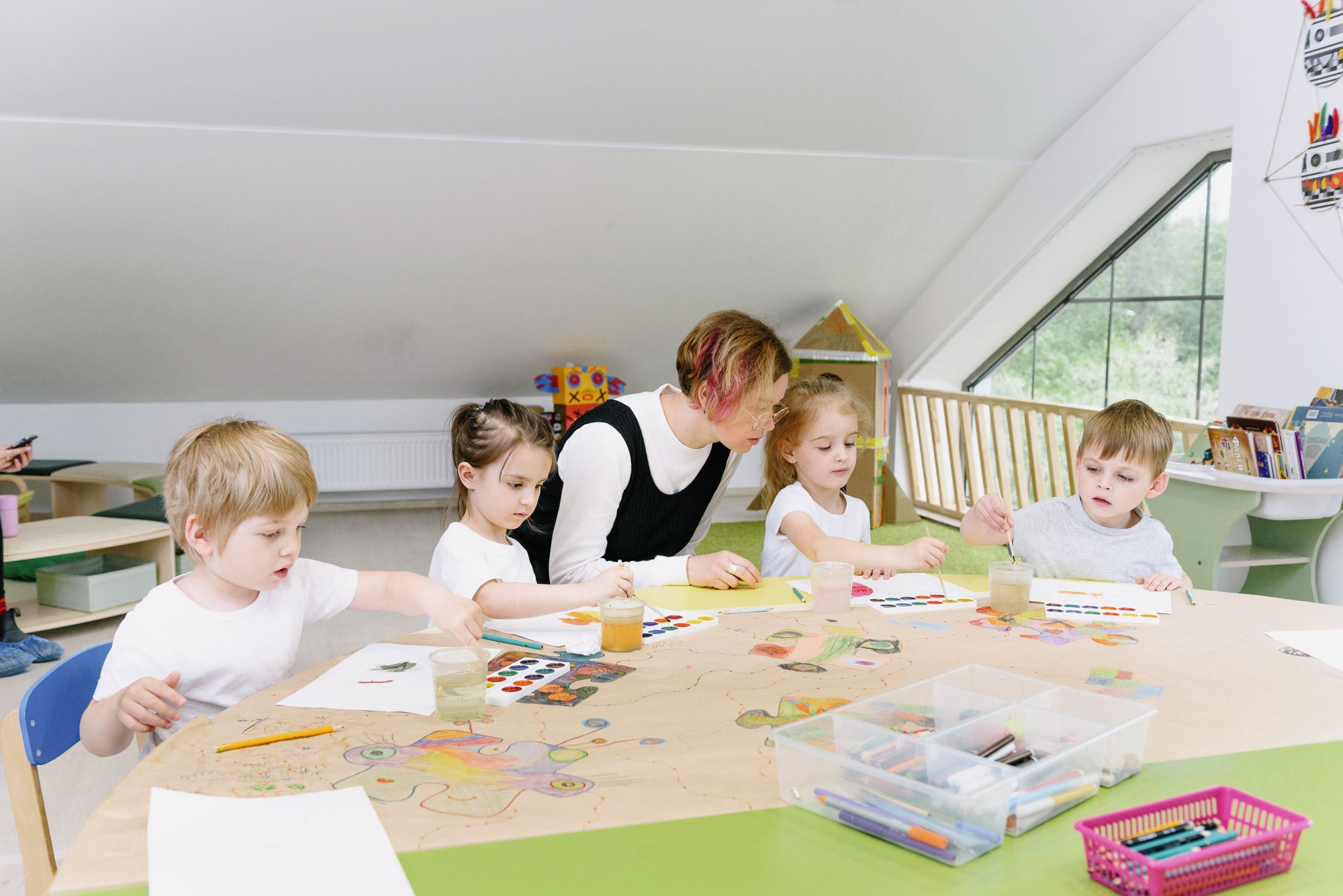 A teacher leaning over a low table where four children are drawing, paints and paper spread out in front of them.