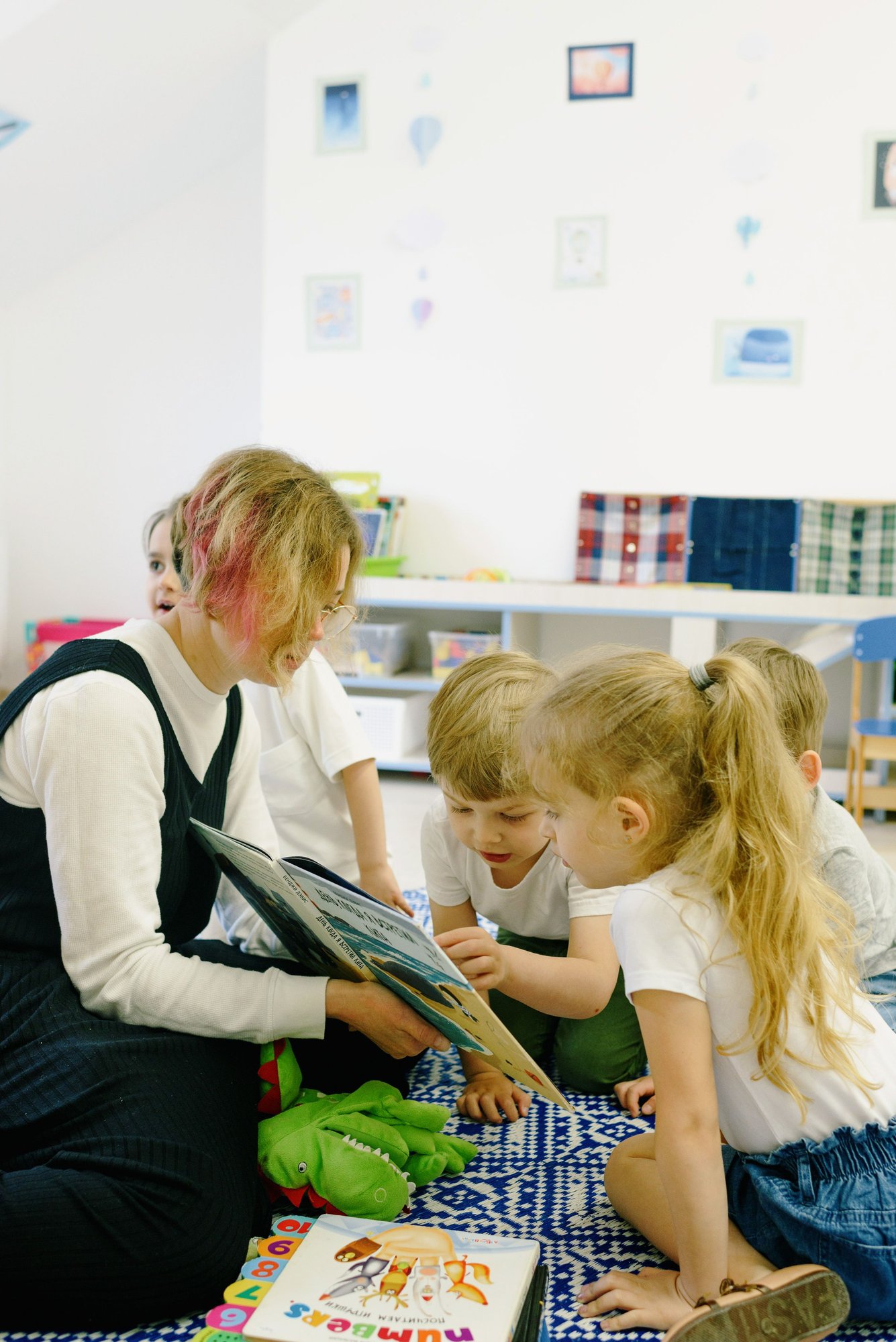 A teacher holding a picture book open for three small children who lean in to look.