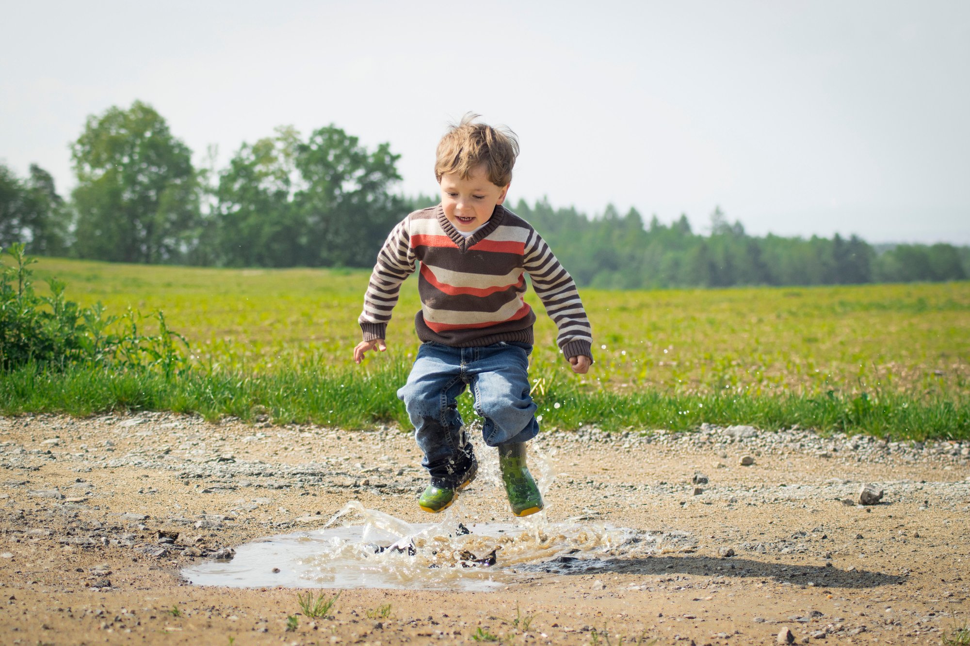 A child in a striped sweater jumping in a field, arms out, trees behind.