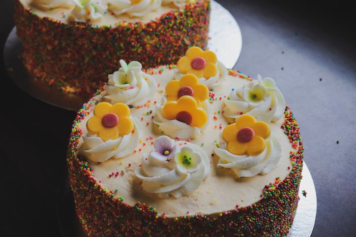 A round cake piped with cream rosettes and yellow sugar flowers, rainbow sprinkles around the base.