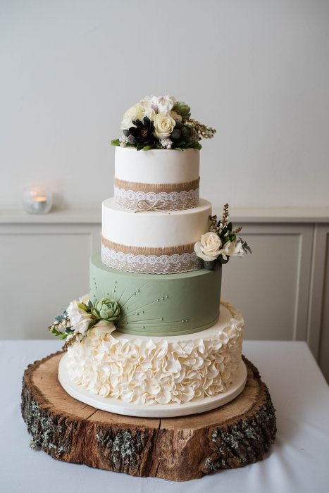 A three-tier wedding cake in cream and sage on a slice of tree trunk, small white flowers tucked between the tiers.