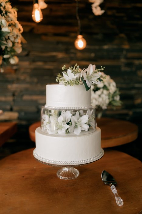 A two-tier white wedding cake with white lilies and eucalyptus on a wooden stand.