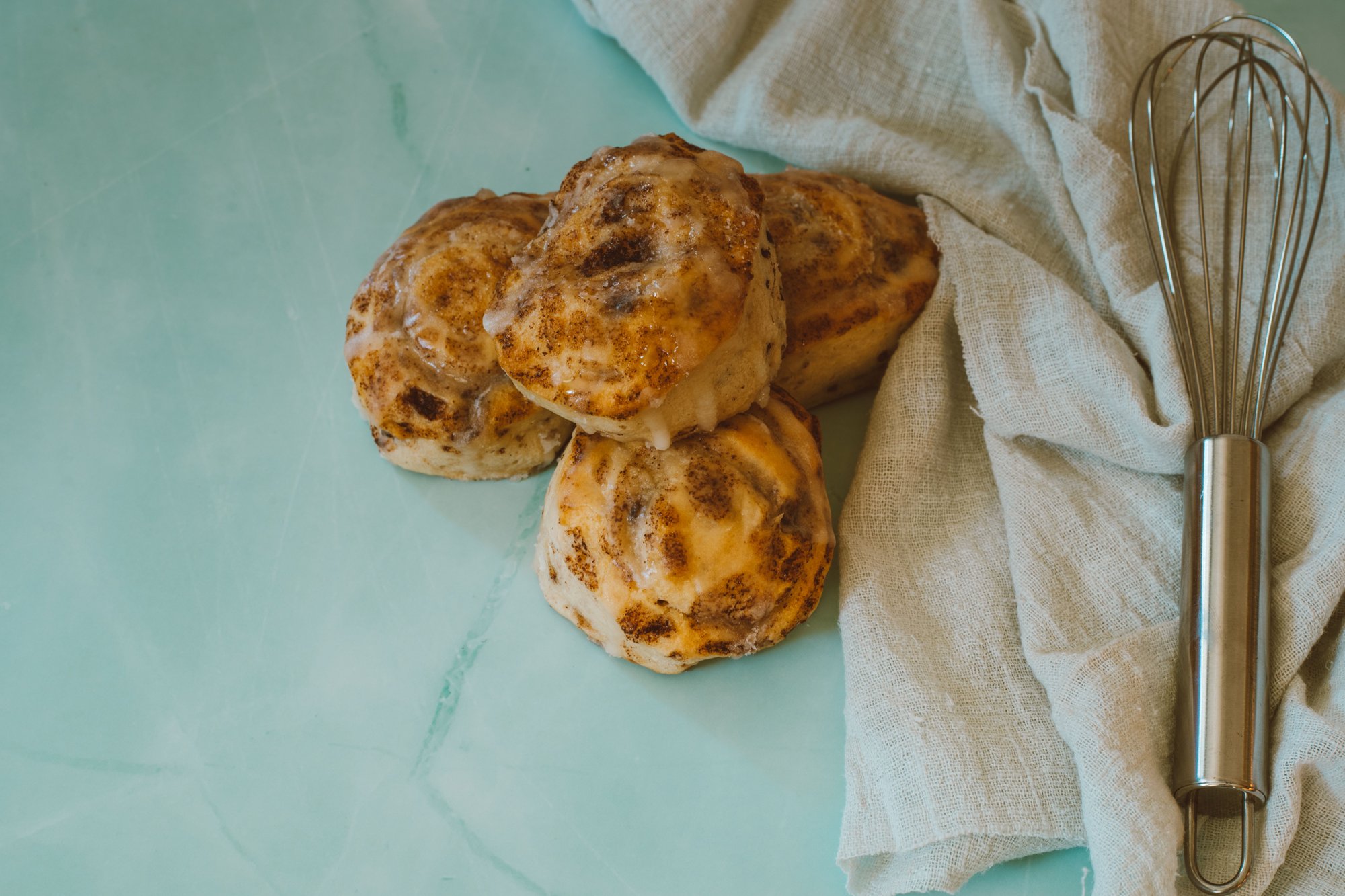 Cinnamon rolls on a pale counter with a whisk and a linen cloth beside them, seen from above.