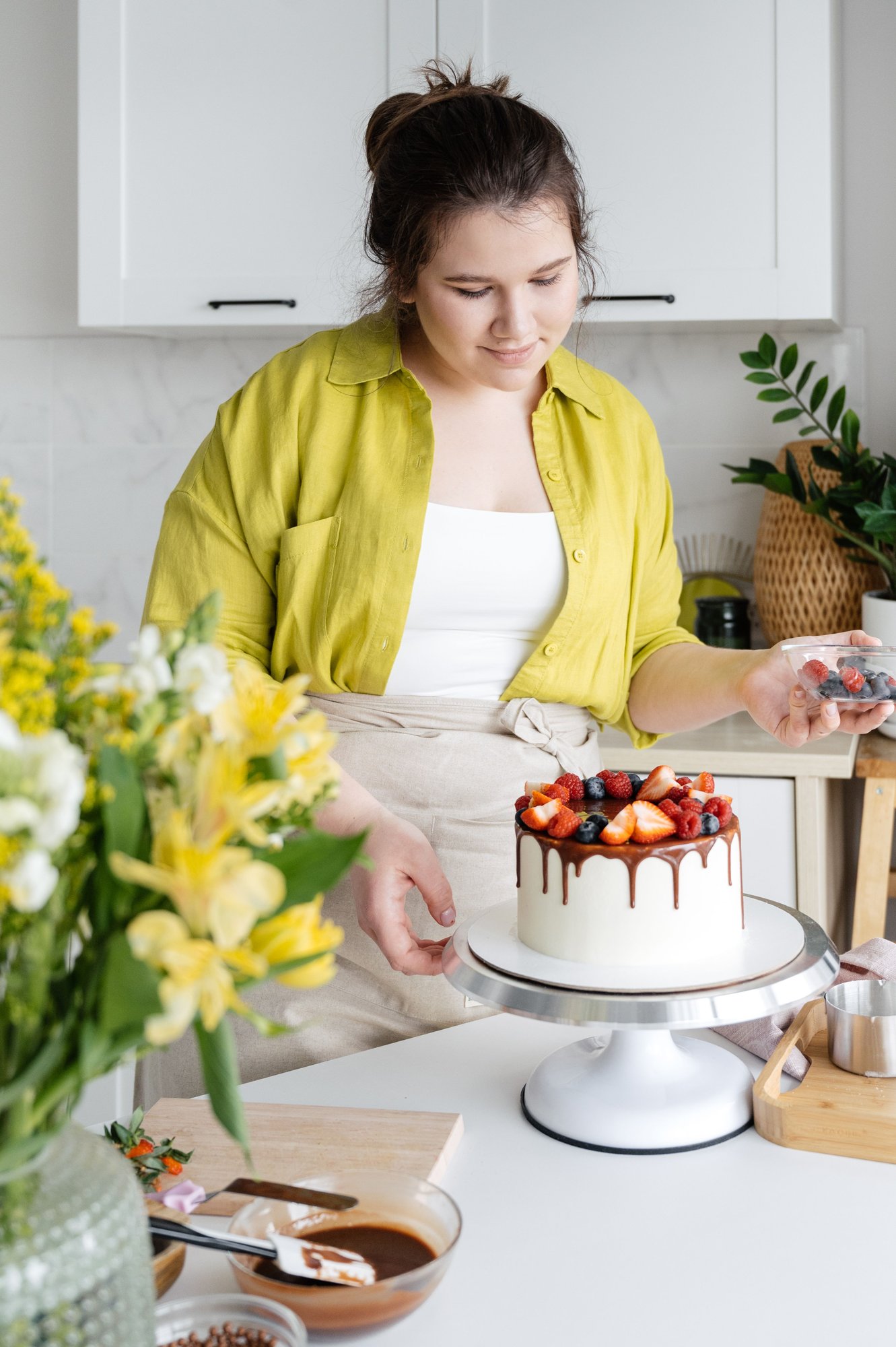A baker in a yellow shirt setting strawberries on top of a finished cake, in a bright kitchen with flowers on the counter.