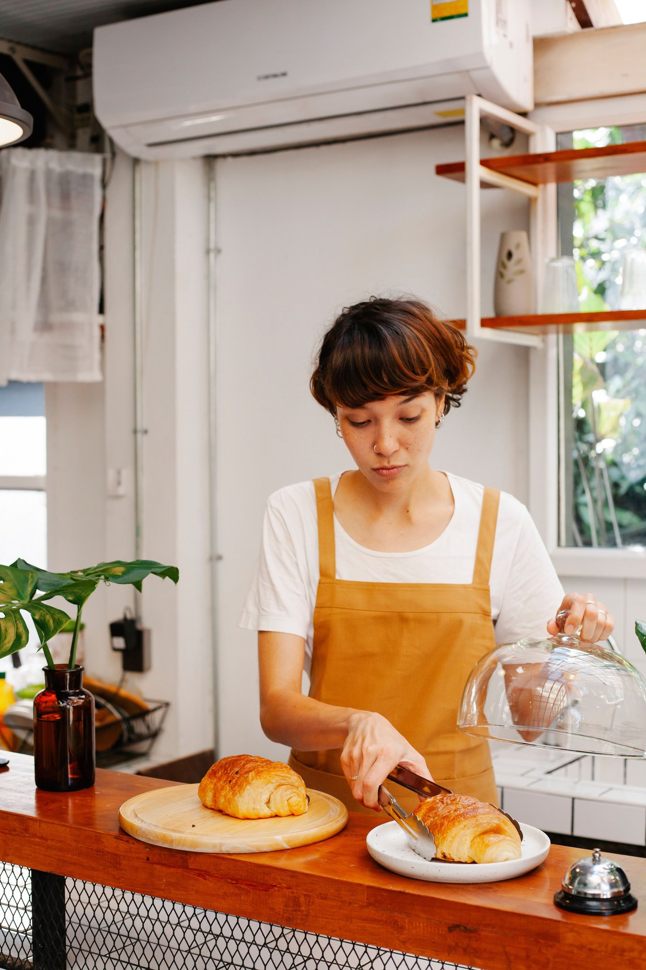 A young baker in an ochre apron setting a loaf down on the counter of a bright shop.