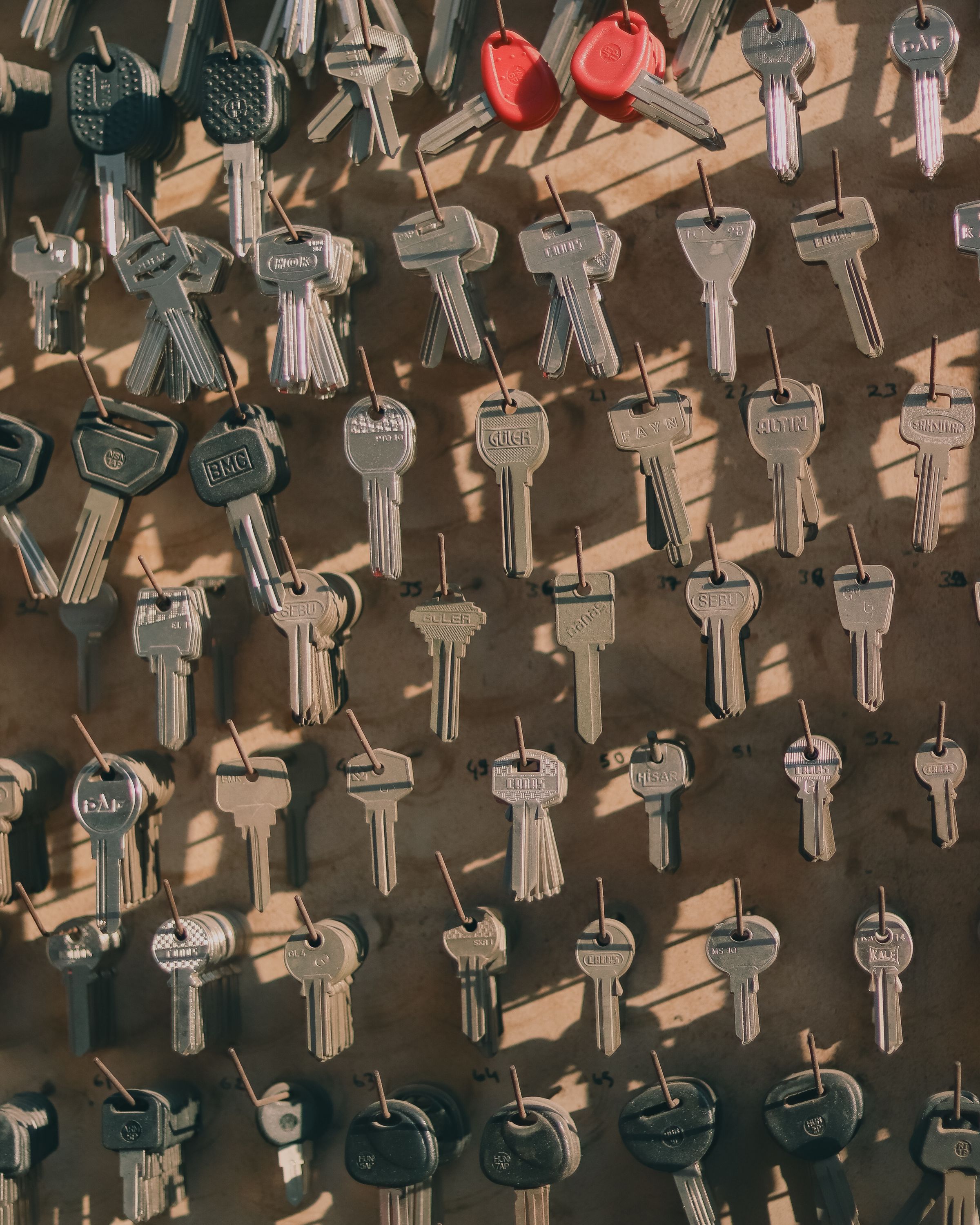 A locksmith’s key board: dozens of cut keys and blanks on numbered hooks over tan pegboard, two red-headed car keys near the top.