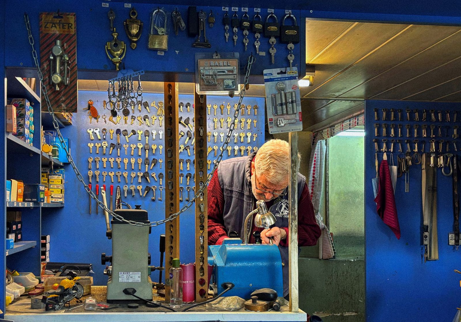 A locksmith with white hair and glasses bent over a key-cutting machine, walls of key blanks and padlocks behind him.