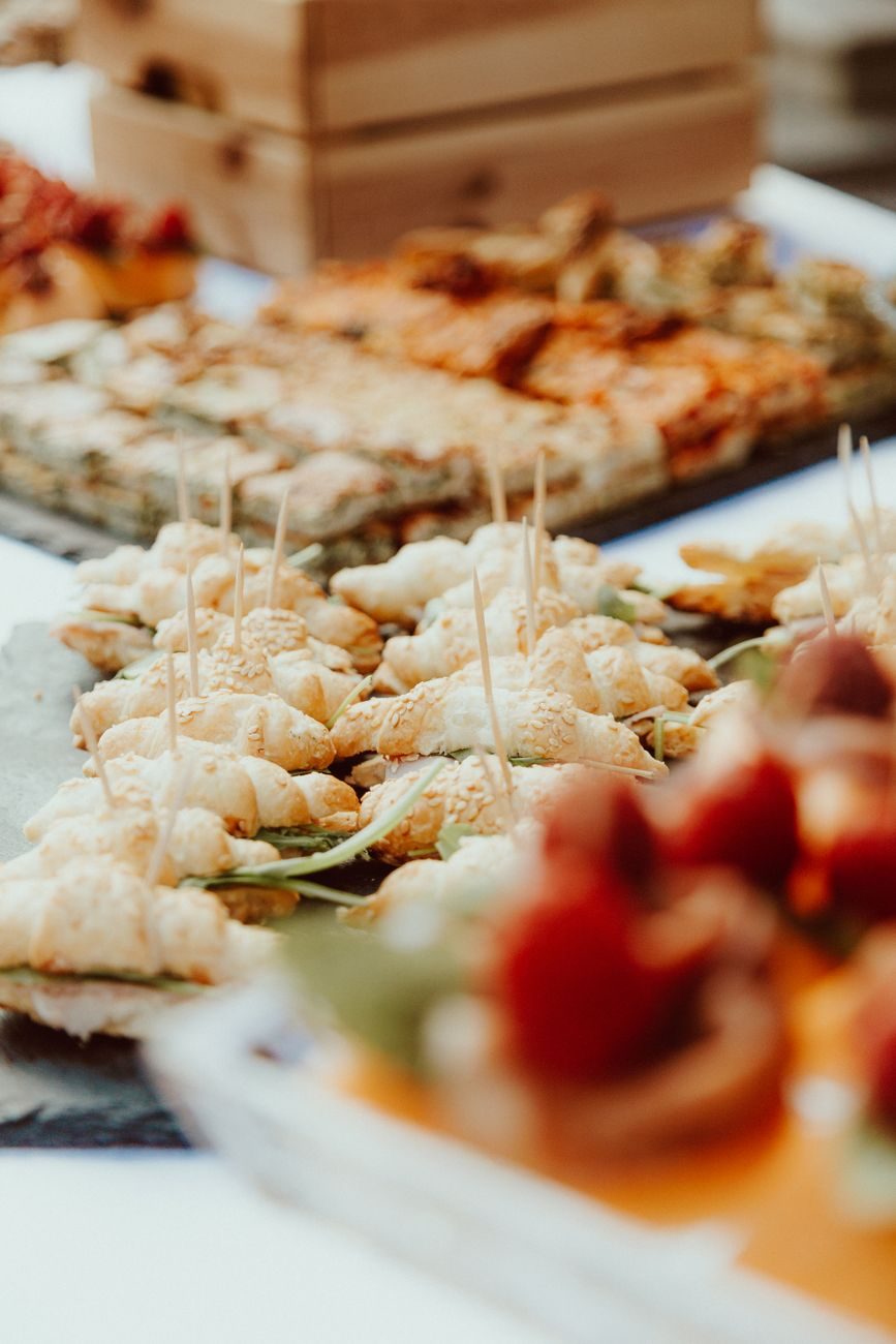 Trays of sesame-topped croissant bites, herbed focaccia squares and strawberry skewers set out on a catering table in warm daylight
