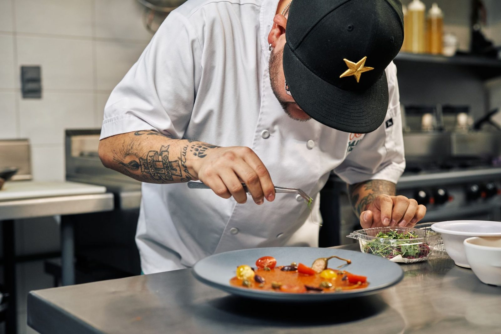 A chef in kitchen whites leaning over a plate, placing microgreens onto a tomato dish with tweezers