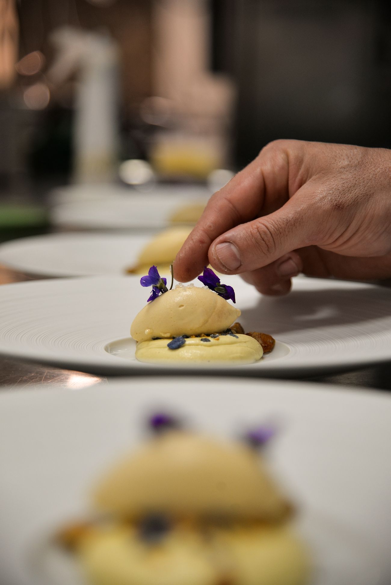 A hand placing an edible flower onto a finished plate, the next plate waiting behind it