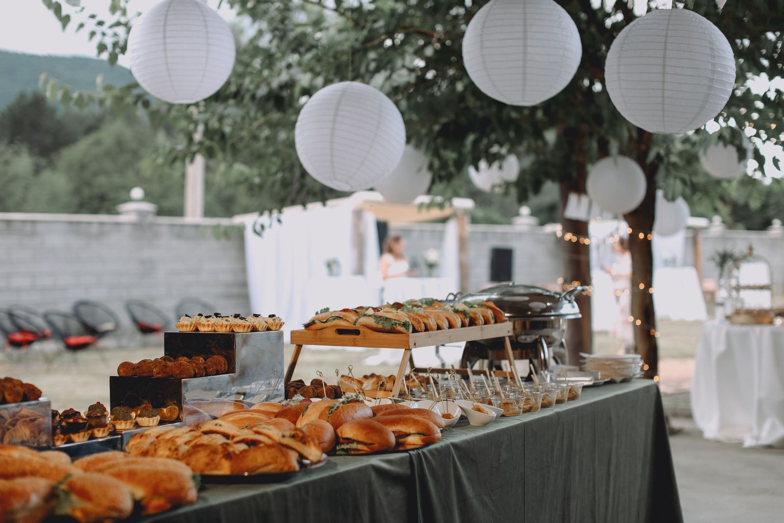 A long buffet laid out outdoors under paper lanterns, trays set down its whole length