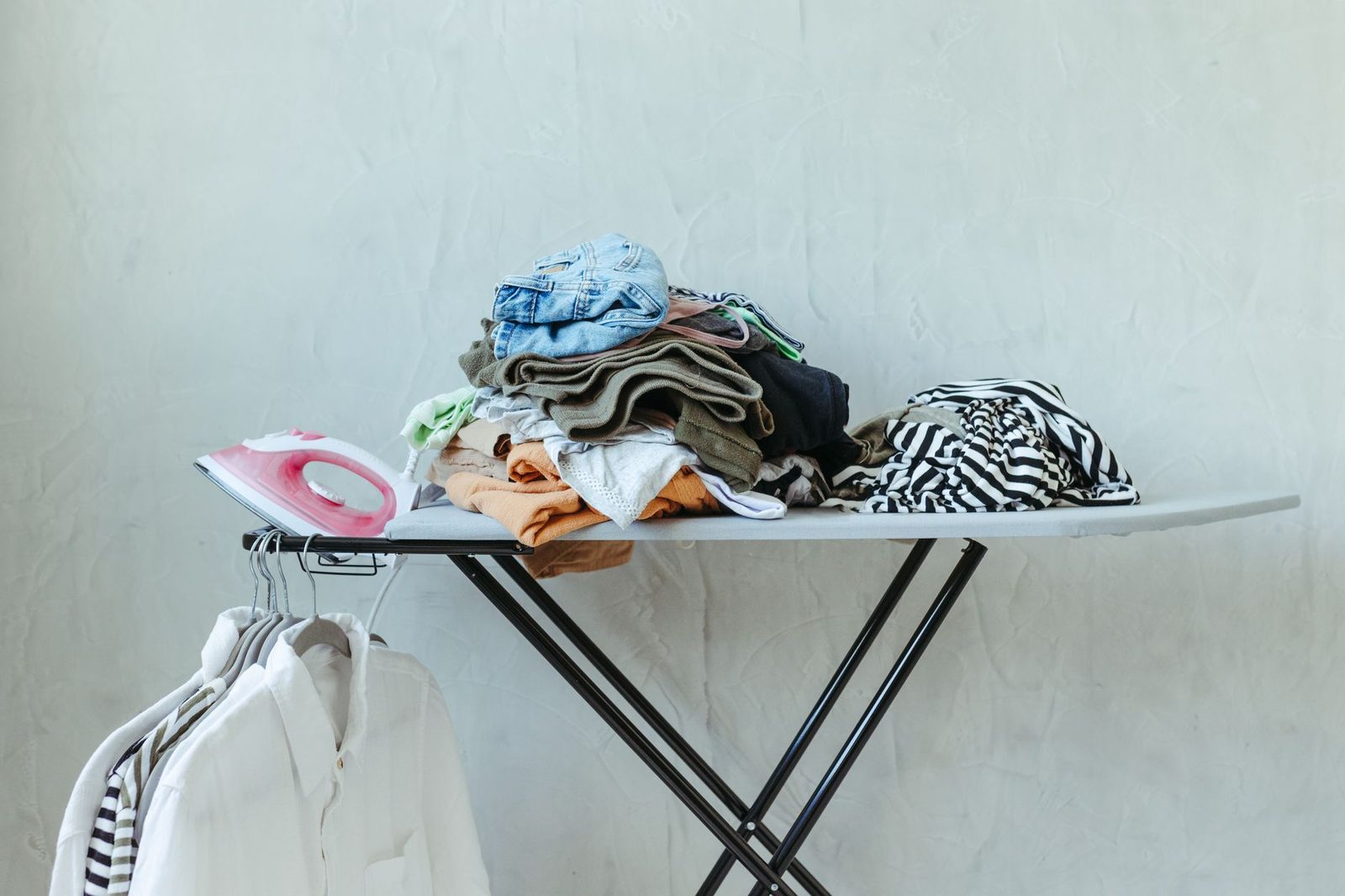 An ironing board holding a stack of shirts, jeans and knitwear beside an iron, with shirts on hangers behind
