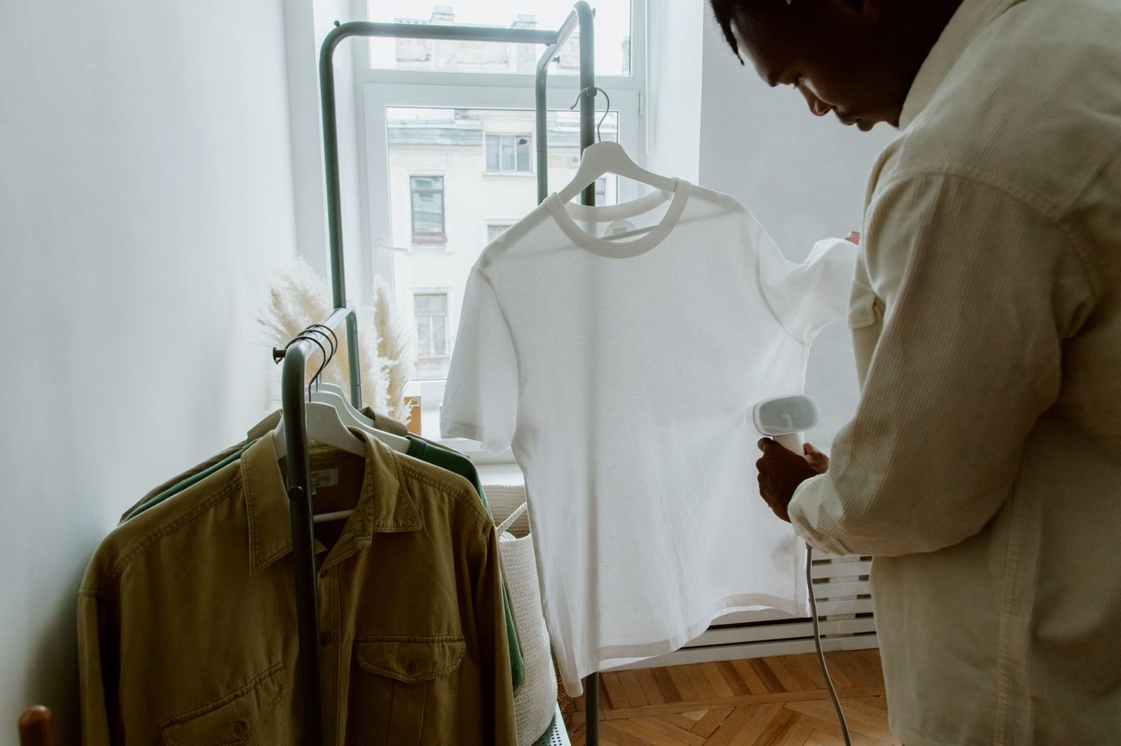 A man steaming a white shirt hanging on a rail beside a bright window