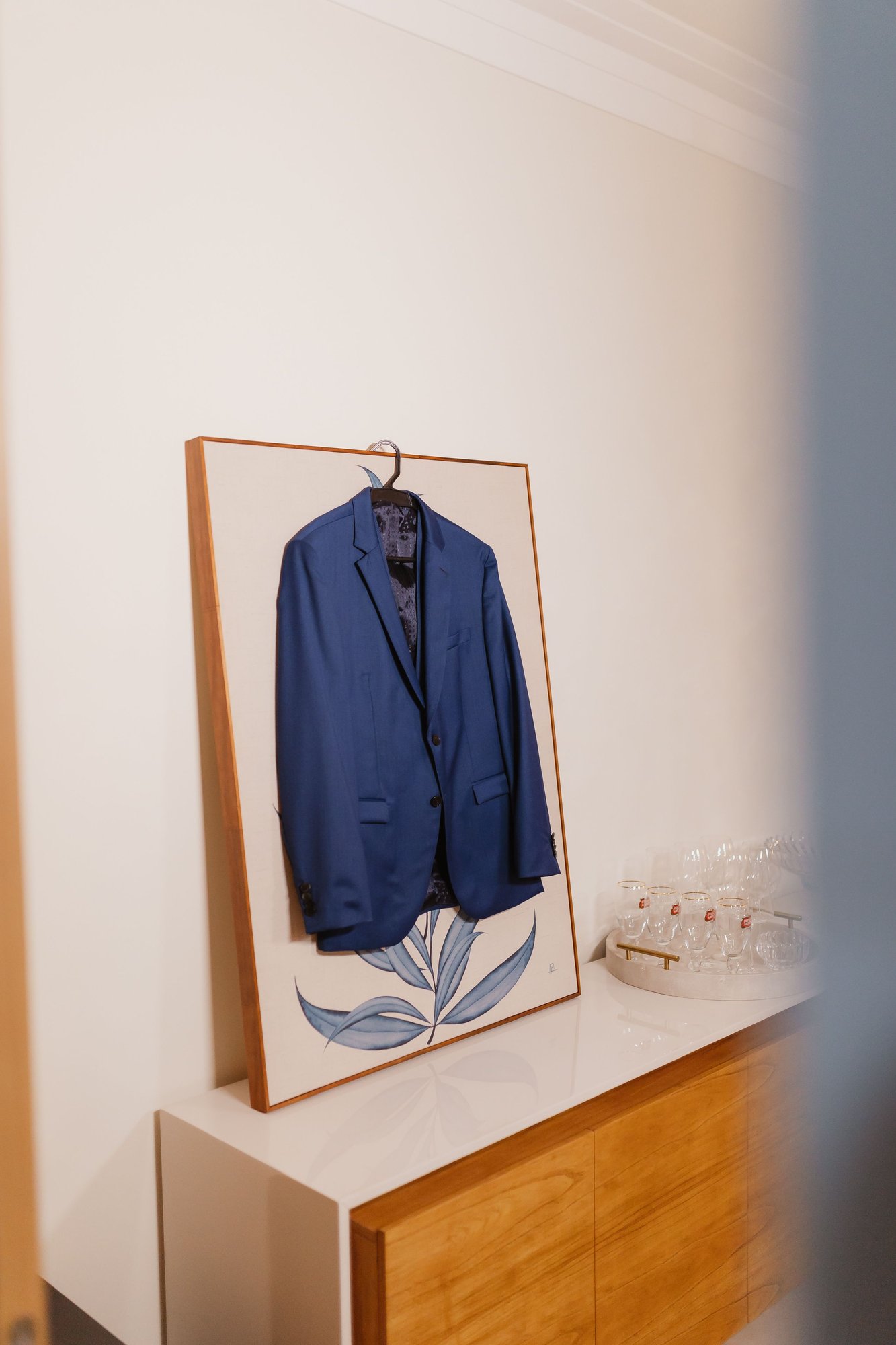 A blue suit jacket on a wooden hanger, hung against a pale wall.