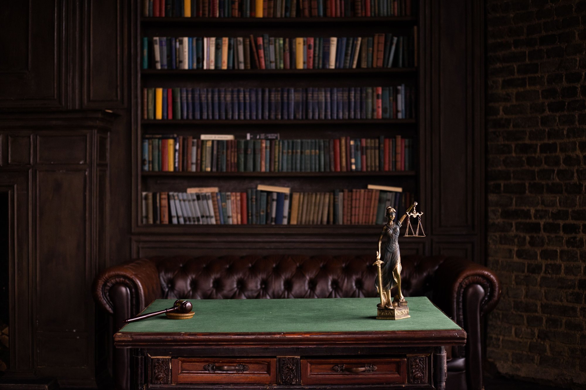 A study with dark wooden shelves of books, a leather sofa, and a green-topped desk holding a gavel and a figure of Justice.