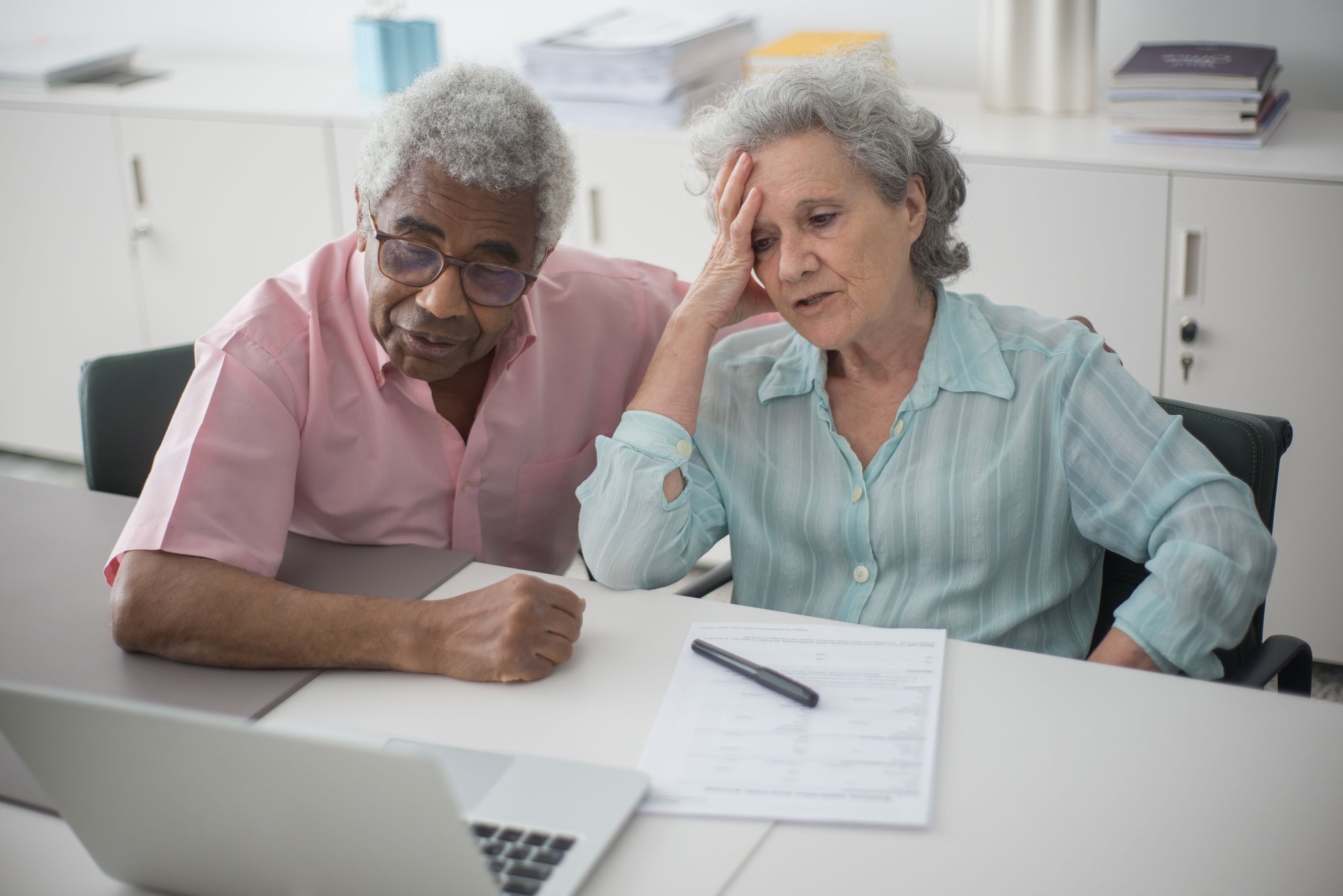 An older couple at a table with a laptop and paperwork; she rests her head in her hand.