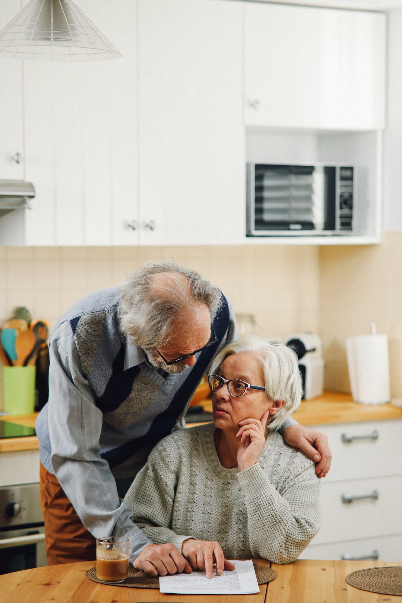 An older man leaning over the shoulder of an older woman at a kitchen table, both looking at paperwork.