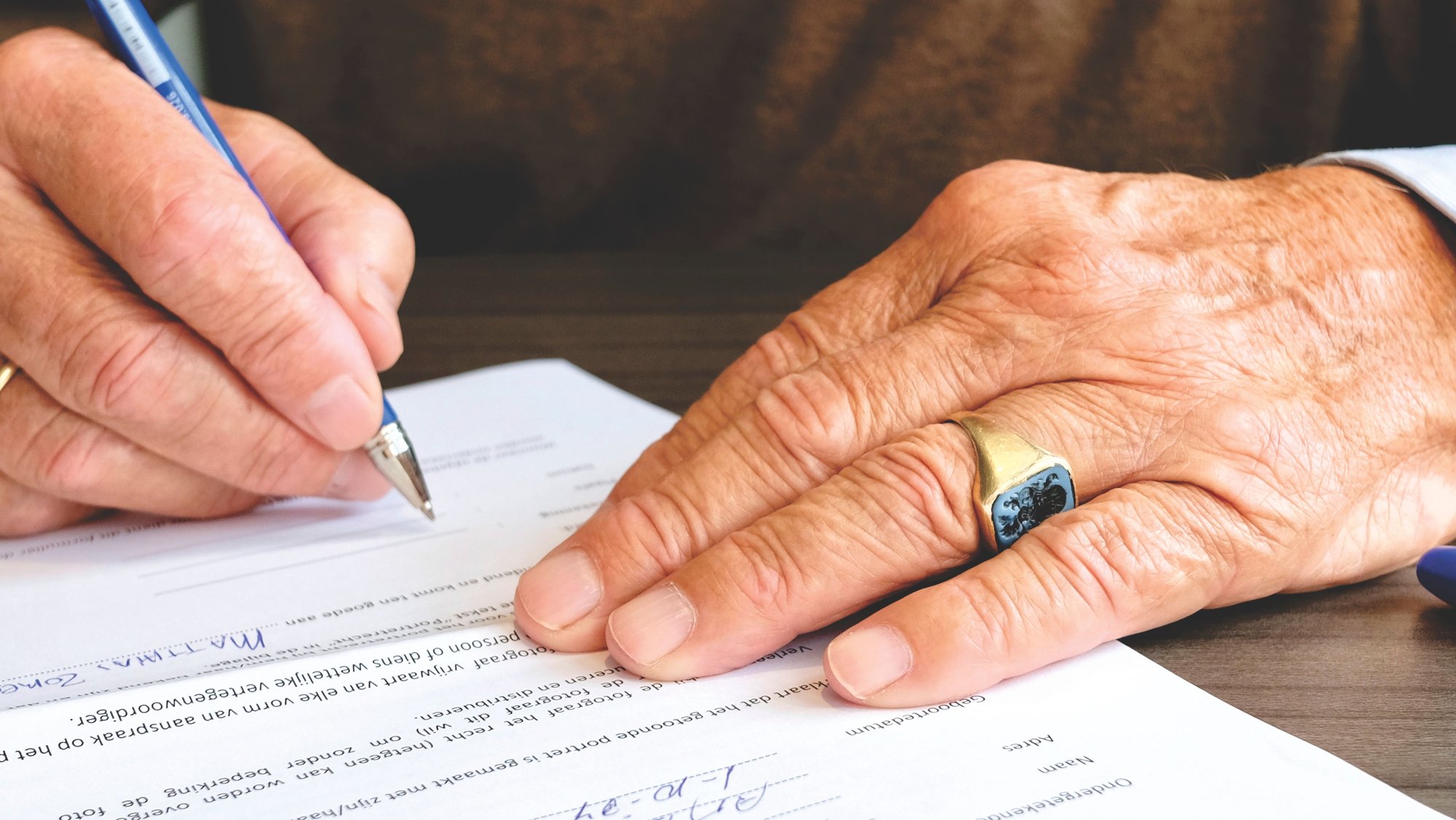 An older hand with a ring signing a document with a ballpoint pen.