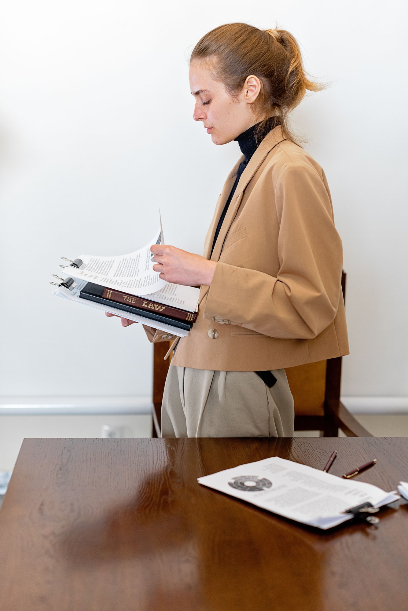 A woman in a camel coat standing at a desk, reading a file.