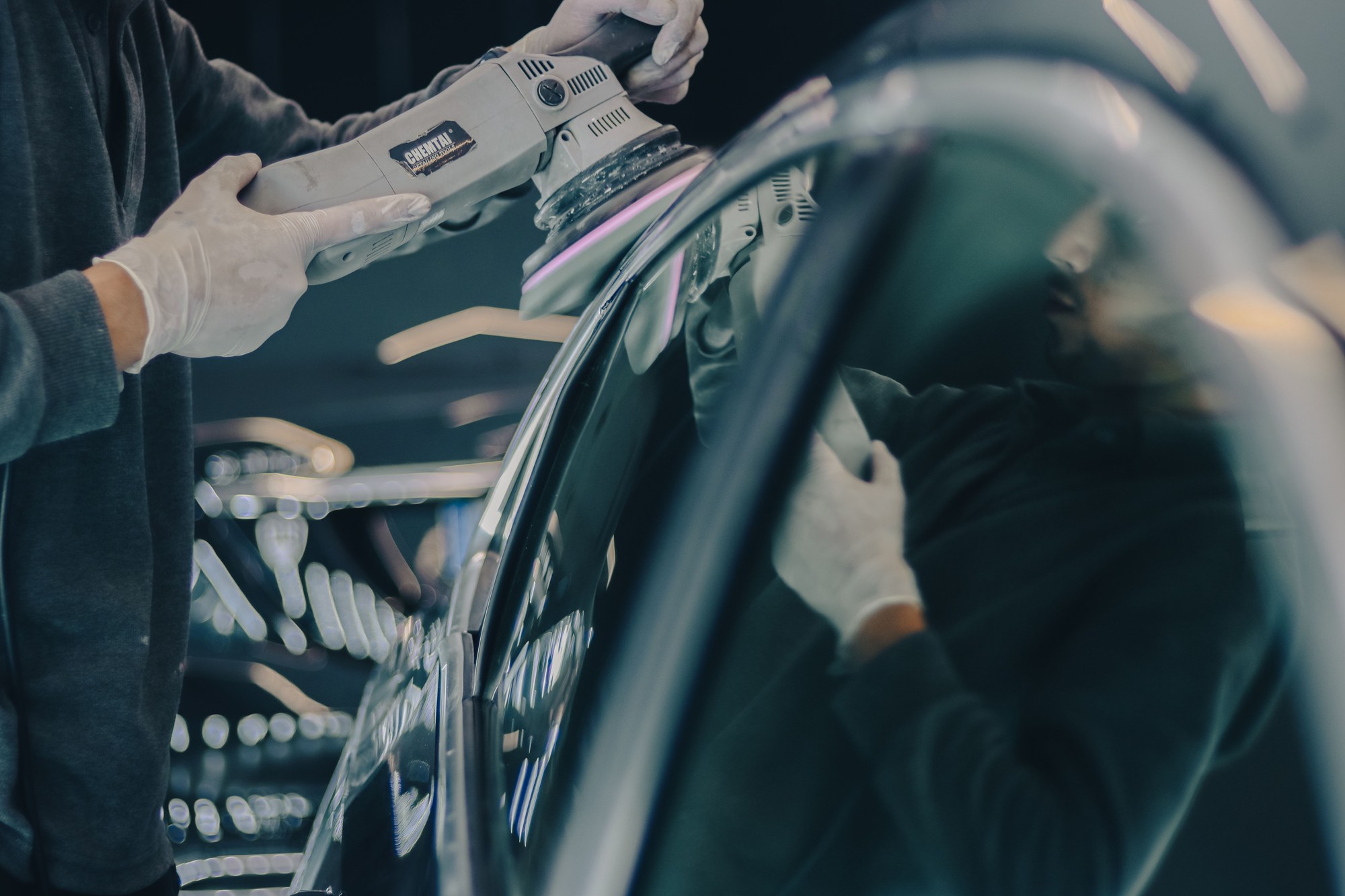 A detailer’s gloved hands guiding a dual-action polisher along the edge of a dark car panel in the shop.