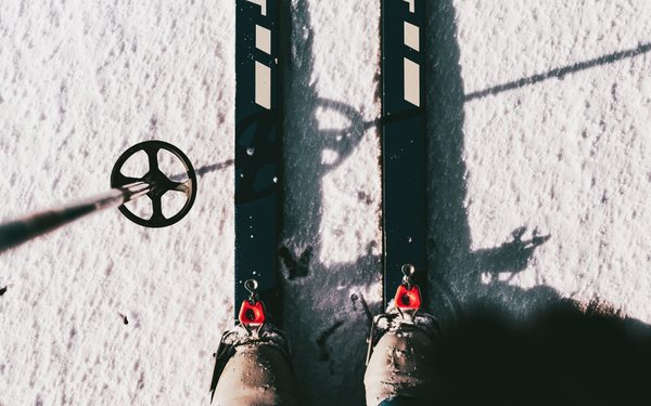 Looking straight down at a skier&rsquo;s boots clipped into their bindings on fresh snow, one pole planted beside them.
