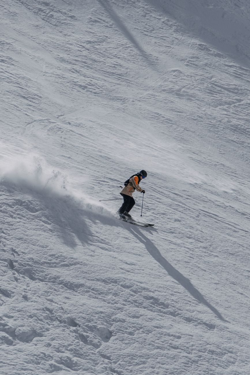A skier carving down a wide, sunlit open slope with a plume of snow trailing behind them.