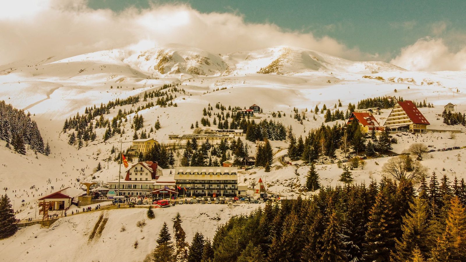 Aerial view of a snowed-in mountain village: hotels and a lift base below, groomed pistes climbing the ridge above, spruce trees in the foreground.