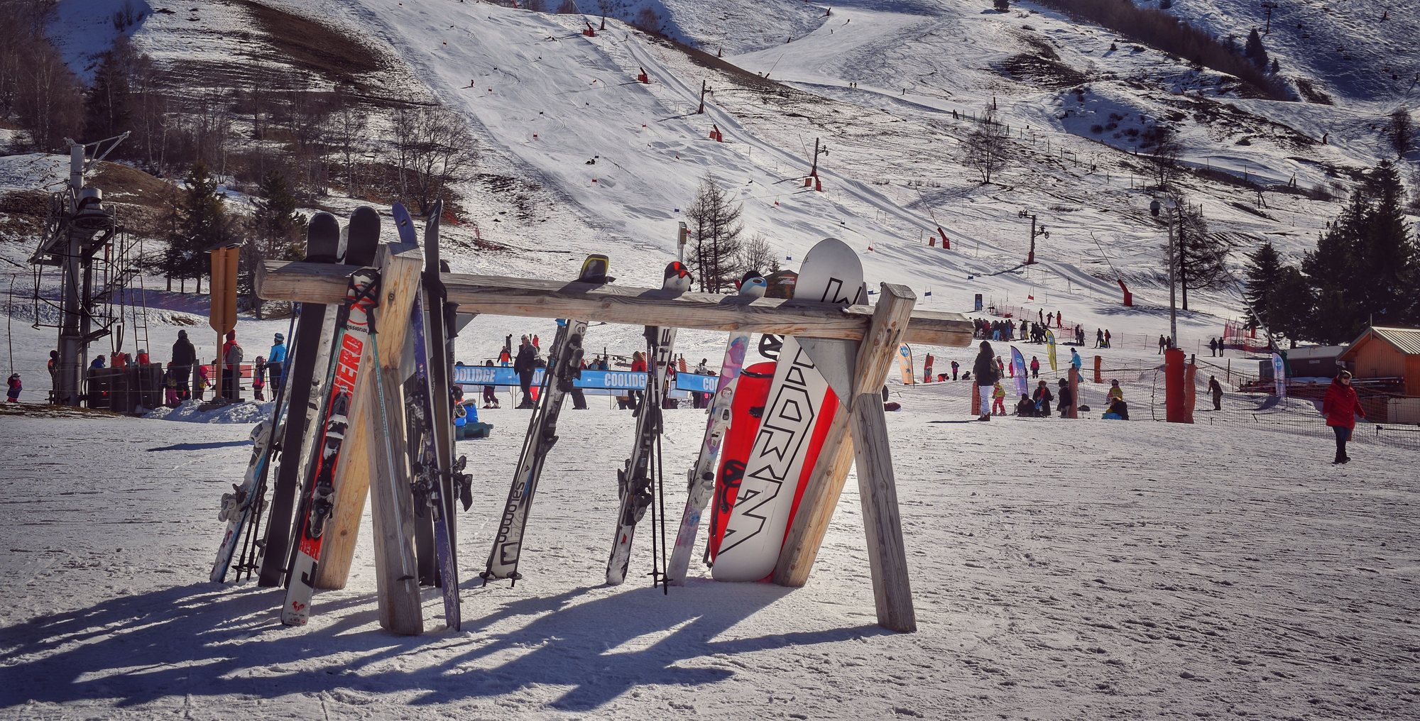 Skis and snowboards standing in a wooden rack at the base of the slopes, skiers and a lift line in the sun behind.