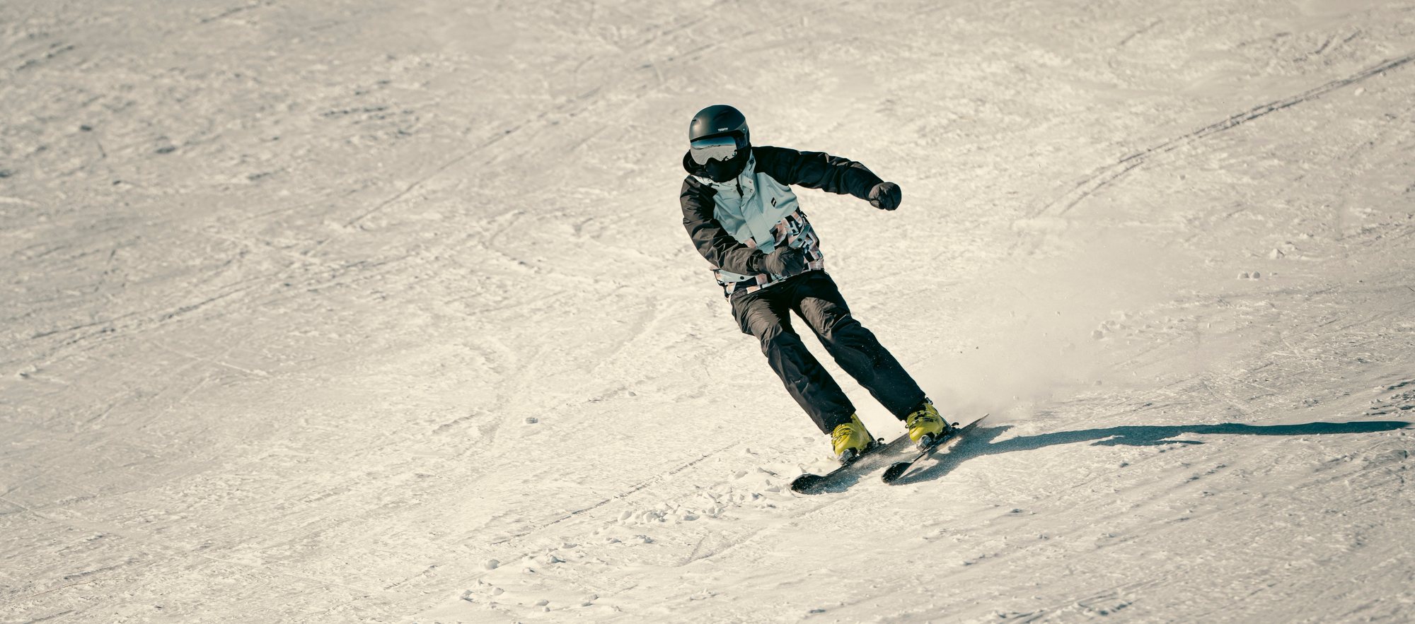 A skier in a black helmet carving a turn down a wide, tracked-out slope in flat afternoon light.