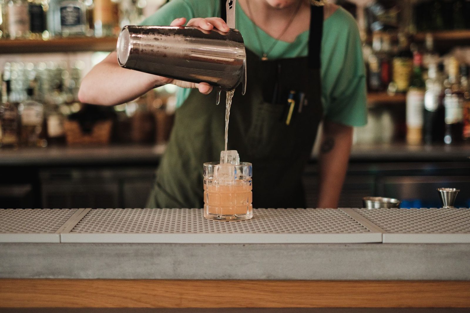 A bartender straining a pale orange cocktail from a shaker over a single large ice cube in a heavy tumbler.