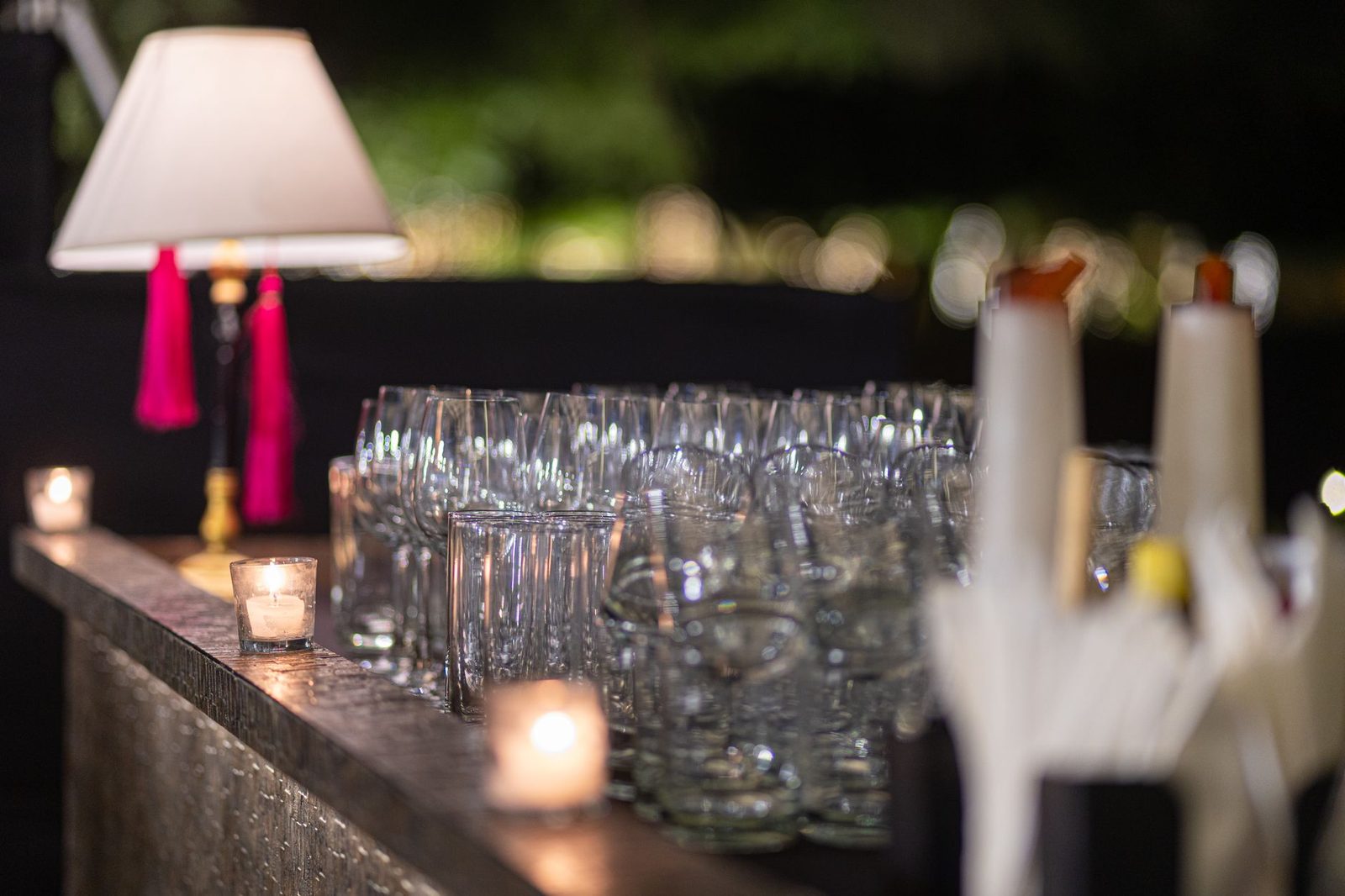 Rows of empty wine glasses and tumblers along a dark bar at night, votive candles burning between them.