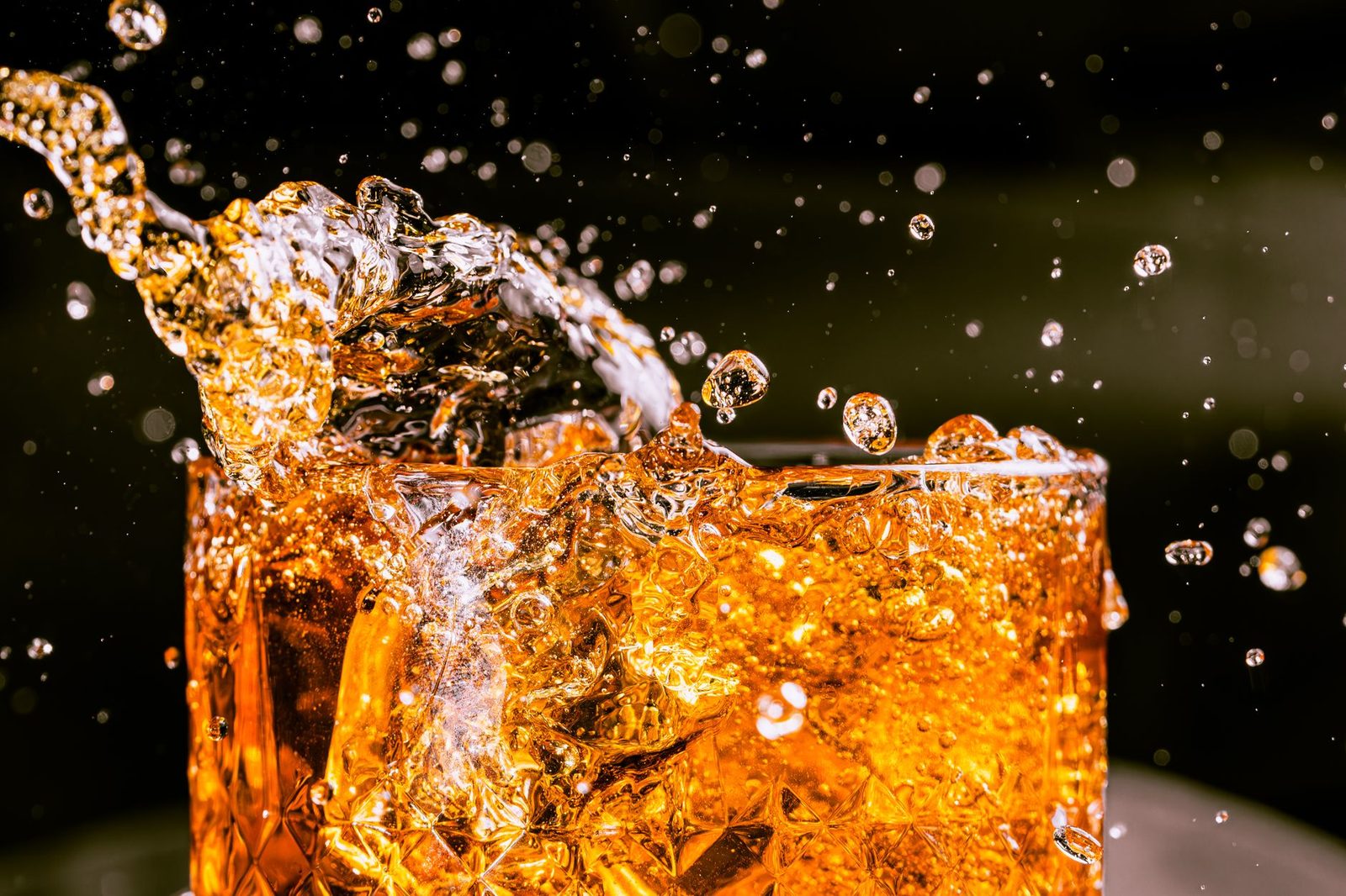 Whiskey splashing over cracked ice in a heavy cut-glass tumbler, gold against a near-black background.