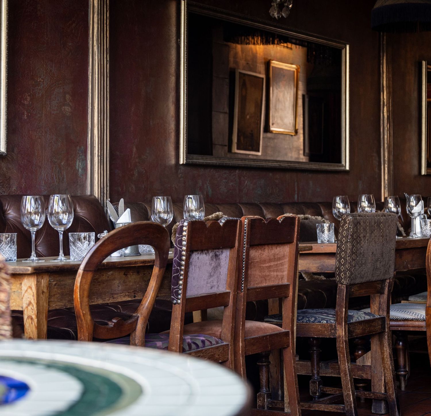 A long table set with wine glasses along a leather banquette, antique chairs and gilt frames on a deep burgundy wall.
