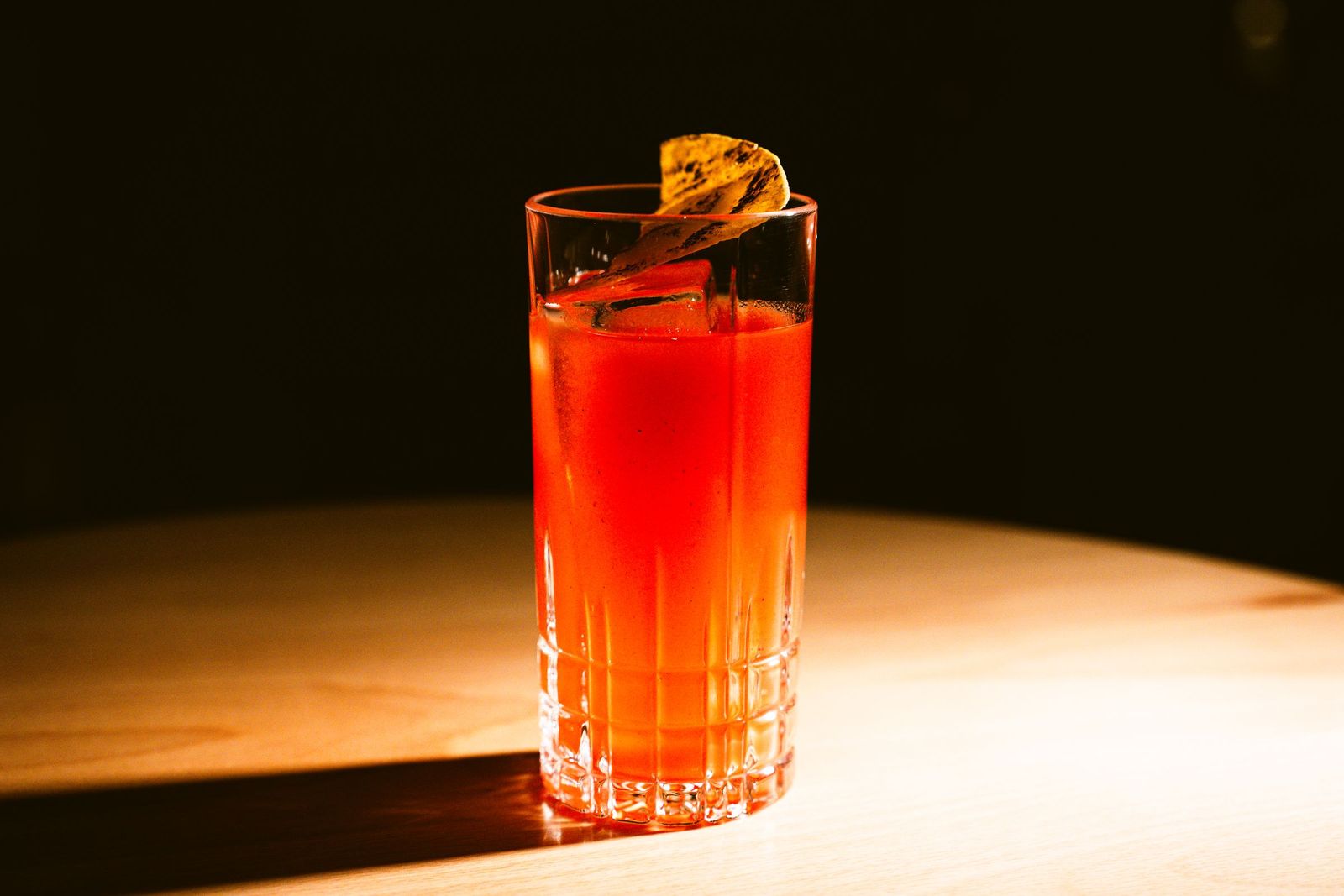 A tall red cocktail with a citrus twist, lit against a dark wooden bar top