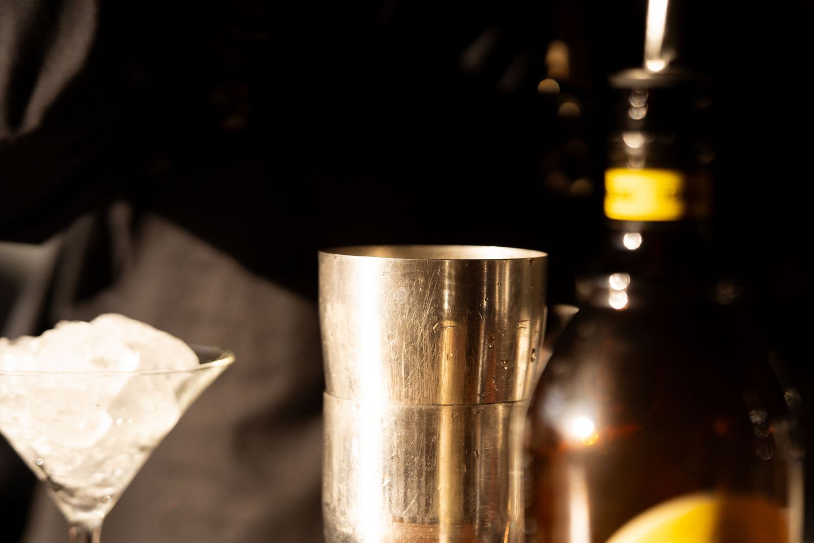 A cocktail shaker and ice under the bar light, bottles standing beyond