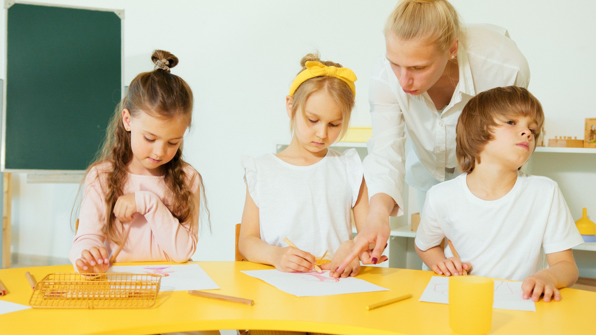 A teacher crouched beside three children drawing at a low table in a bright classroom.